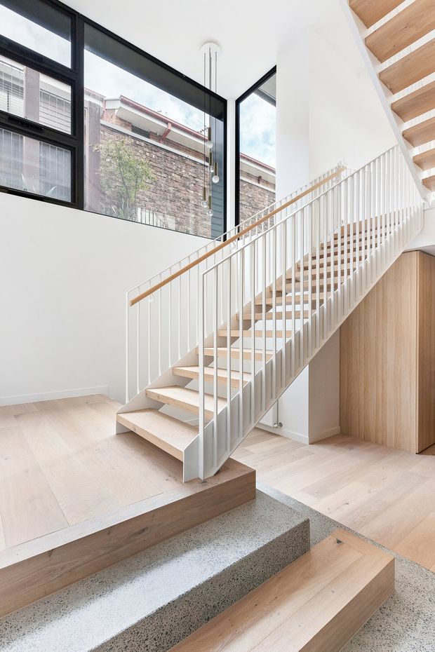 Bright contemporary stairway interior with a pale timber open-riser staircase, slender white vertical balustrades, and a timber handrail. Large black-framed windows provide abundant natural light and overlook a neighbouring brick wall. Pale timber flooring, white walls, and a terrazzo-like stepped landing create a clean, refined architectural composition. Built-in timber joinery is visible beneath the stair.