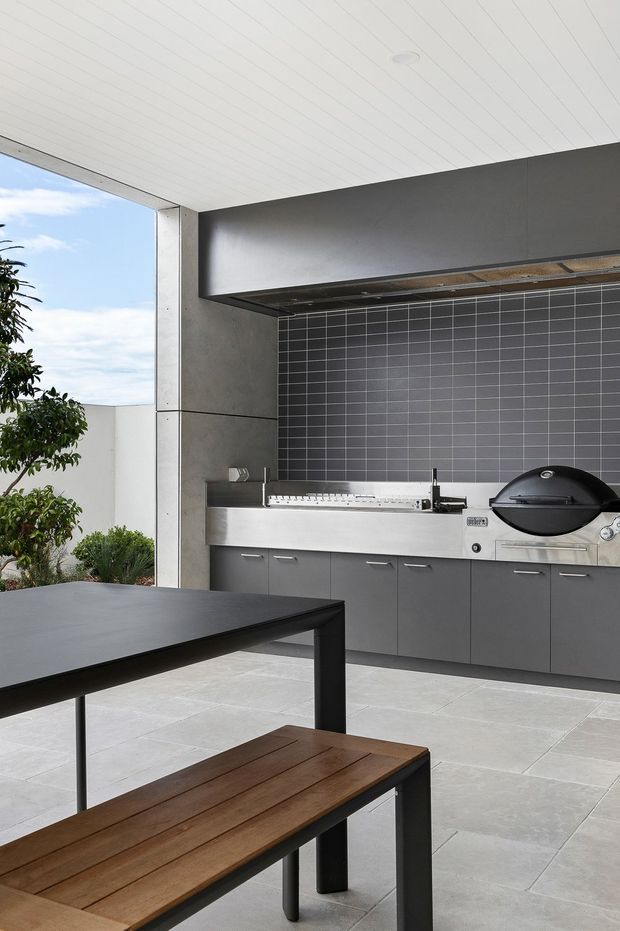 A covered outdoor entertaining area with a built-in stainless-steel barbecue kitchen, dark tiled splashback, grey cabinetry, and a black grill. A large dark dining table with a timber bench occupies the foreground. Pale tiled flooring, concrete-look walls, and a white panelled ceiling create a contemporary, structured setting, with garden greenery visible at the open side.