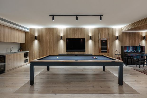 A refined recreational room centered on a full-size black pool table, with a wall-mounted television, warm vertical timber wall panelling, integrated cabinetry and kitchenette along the left wall, and a piano in the corner. Linear track lighting and concealed wall-wash lighting create a warm, contemporary atmosphere.