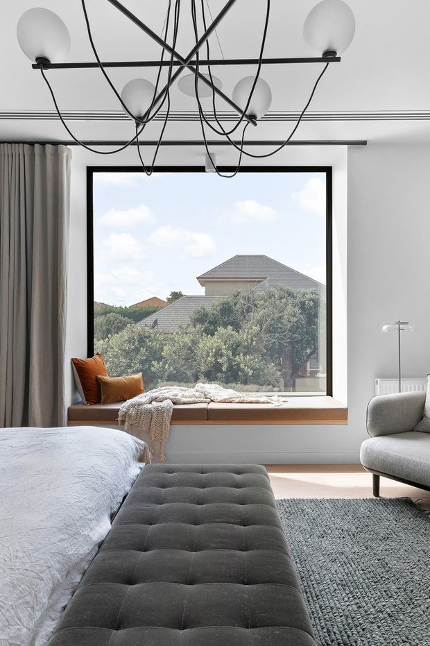 A bright, contemporary bedroom with a large black-framed picture window and built-in timber window seat. The room includes a partially visible bed with pale bedding, a long dark upholstered button-tufted bench, a grey armchair, textured grey rug, full-height grey curtains, and a sculptural black pendant light with exposed looping cords. Trees and neighbouring rooftops are visible through the window.