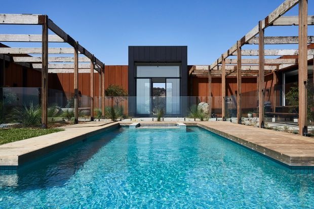 A contemporary courtyard centred on a long rectangular swimming pool, with timber pergola structures and transparent glass balustrades on both sides. Rust-coloured cladding forms the courtyard walls, while a dark-framed glazed pavilion or entry volume anchors the far end. Pale stone or concrete paving surrounds the pool, with sparse planted landscaping and a clear blue sky creating a bright outdoor setting.