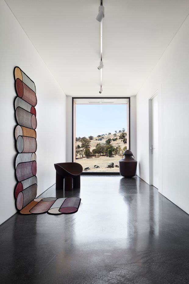 Minimalist, gallery-like interior hallway or entry space with white walls and ceiling, polished dark grey concrete flooring, a linear track light, and a large glazed opening framing a dry rural landscape. Sculptural rounded wall artwork and matching floor pieces add muted burgundy, mauve, beige, and grey accents. A dark sculptural chair and large ceramic vessel sit near the window, while a white door opens on the right.