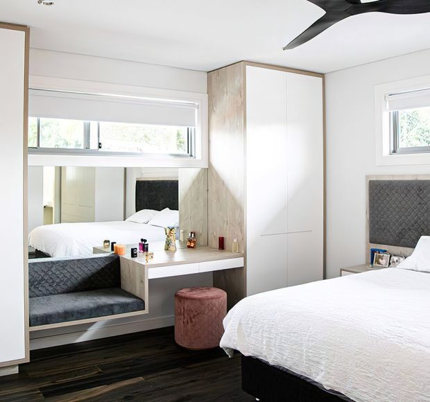 Bright contemporary bedroom with a white upholstered bed, dark timber flooring, built-in white wardrobes, and a pale timber vanity and dressing nook. A large wall mirror reflects the bed and room, while a textured dark grey upholstered headboard and matching bench add contrast. A black ceiling fan and compact windows complete the clean, functional fit-out.