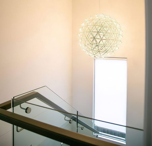 Bright modern stairwell or landing with a glass balustrade, pale peach walls, a tall narrow window with a white roller blind, and a suspended spherical wire-and-glass pendant light. The staircase and glazing create a light, airy architectural interior.