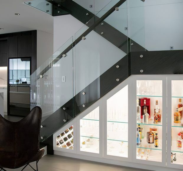 Modern interior view beneath a staircase, featuring a dark timber or wood-look stair stringer and treads, clear glass balustrades with exposed metal fixings, and illuminated built-in glass-fronted cabinetry displaying bottles. A dark leather chair and part of a contemporary kitchen are visible at the left.