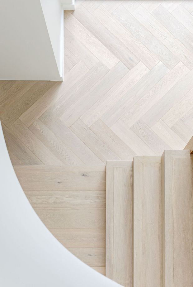 Top-down architectural detail of a light timber staircase and landing. Pale wood flooring is arranged in a herringbone pattern across the landing, transitioning to straight-grain timber stair treads and risers. Smooth white curved walls and crisp white trim frame the stairwell, creating a bright, minimal composition.