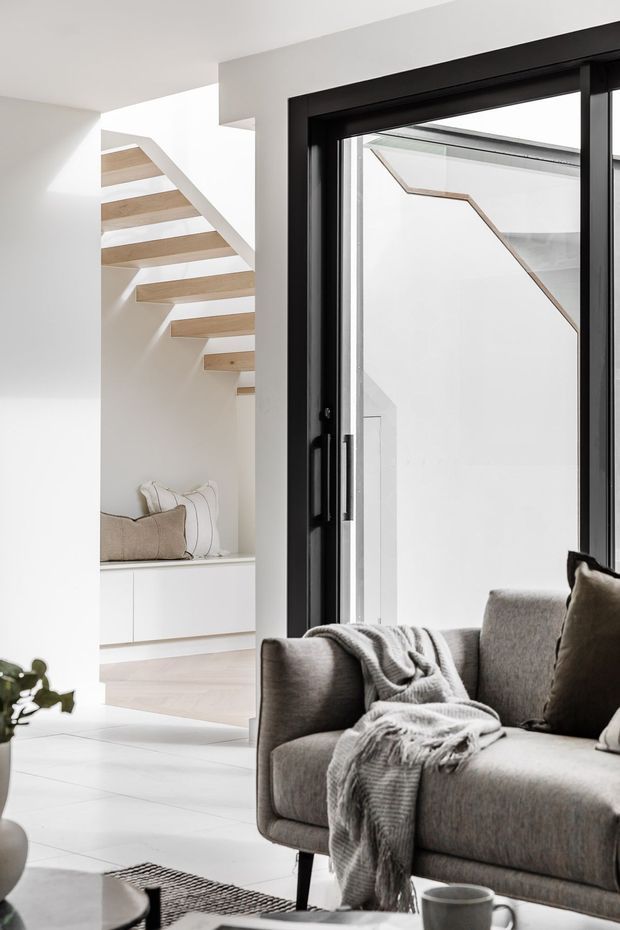 Bright contemporary living area with a grey upholstered sofa in the foreground, layered throw and cushions, and a black-framed glazed door or window. Beyond is a minimalist white hallway featuring an open floating timber staircase, built-in white cabinetry, and a bench with cushions. The space has a restrained neutral palette and strong natural light.