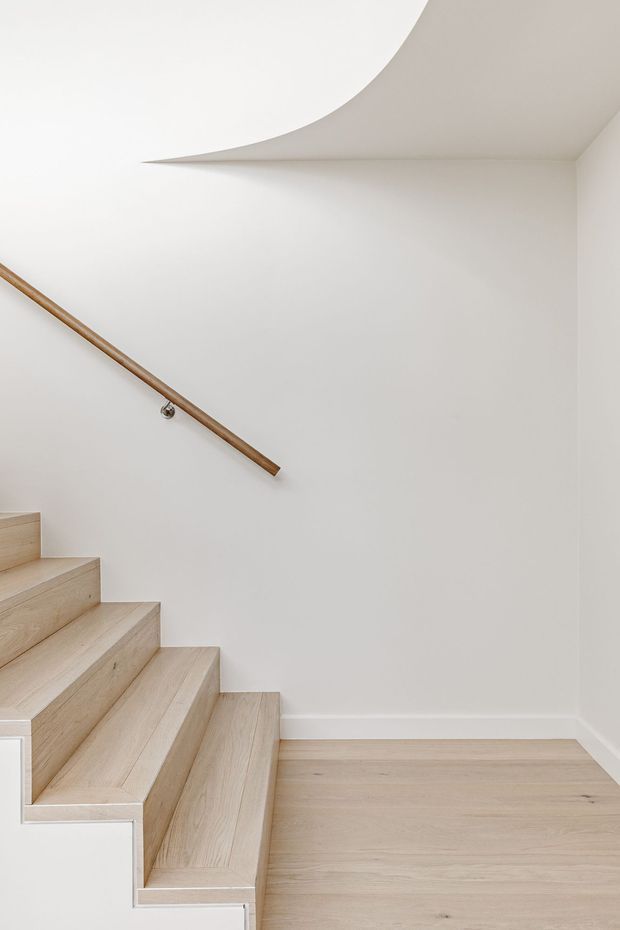 Minimalist interior view of a light-filled stairway with pale timber treads and matching timber flooring. The staircase is enclosed by smooth off-white walls and features a simple cylindrical timber handrail with discreet metal brackets. A sculptural curved ceiling transition creates a distinctive architectural detail.