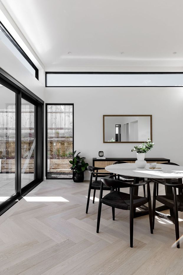 Bright contemporary dining area with a round white dining table and black timber chairs, set against white walls and pale herringbone timber flooring. Large black-framed glazed doors and clerestory windows provide abundant natural light, while a slim dark sideboard, framed mirror, and indoor plants complete the space. A courtyard or garden is visible through the glazing.