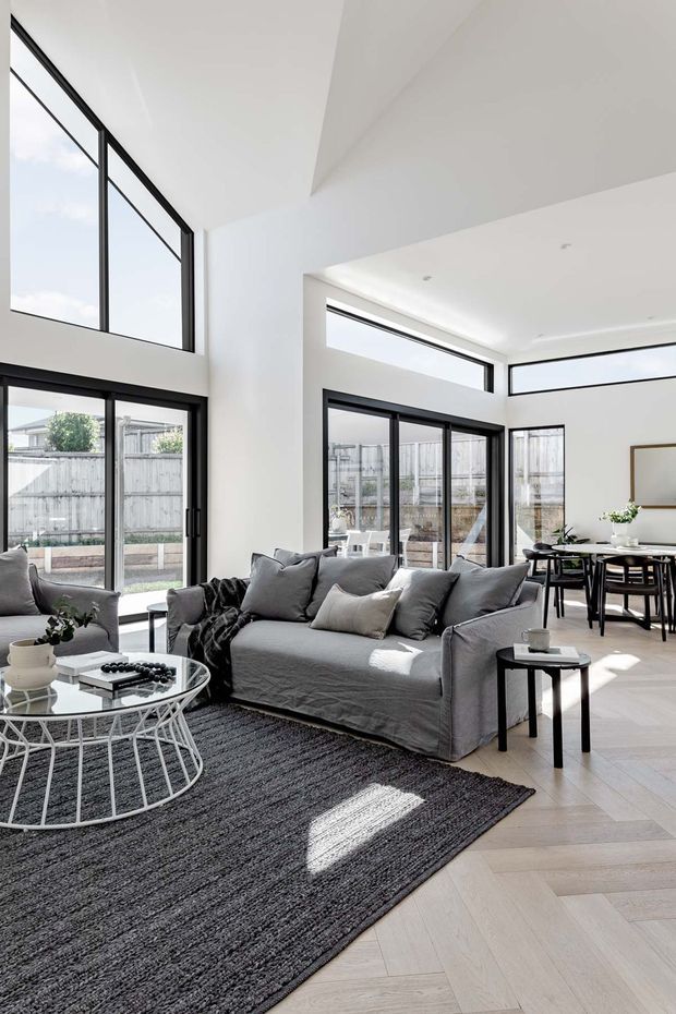 A bright, open-plan living and dining area with a double-height white ceiling, extensive black-framed glazing, and strong natural light. The living zone features a large grey slipcovered sofa, matching armchair, round glass coffee table, dark textured rug, and small black side table. A compact dining setting with a dark table and chairs sits beyond the sofa. Pale herringbone timber flooring and clean white walls create an airy, contemporary interior.
