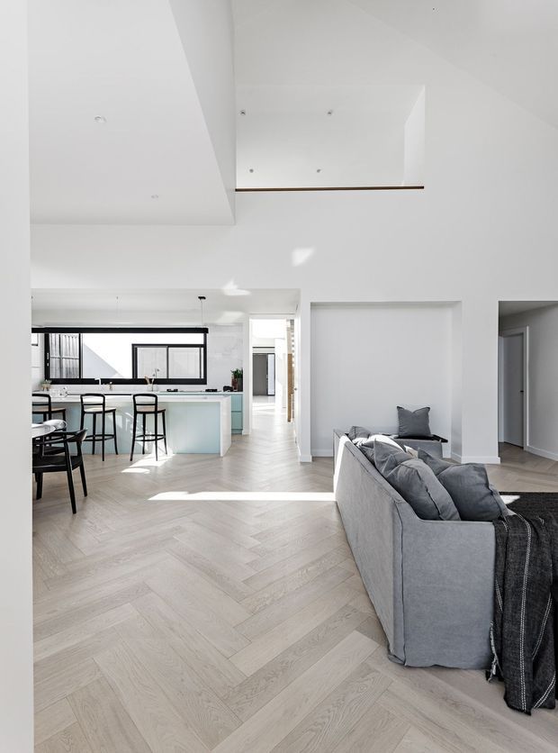Bright open-plan contemporary interior viewed from the living area toward a kitchen and dining zone. The room features pale herringbone timber flooring, white walls and ceilings, a large grey sectional sofa, a light island with pale blue cabinetry, black-framed glazing, and a partial double-height or void-like ceiling detail with a slim timber edge. The palette is predominantly white, light wood, grey, and black, with abundant natural light and a clean refined appearance.
