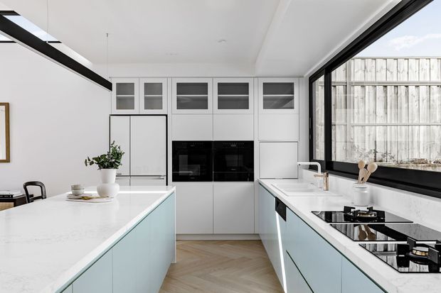 Bright contemporary kitchen with pale blue-grey cabinetry, white stone benchtops, integrated black ovens, a central island, and a wide black-framed window overlooking a timber fence. Herringbone timber flooring and minimal styling create a clean, refined interior.