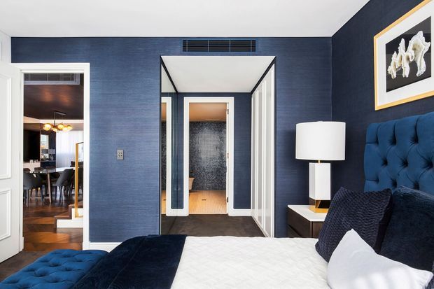 A refined bedroom with deep navy textured walls, a tall tufted navy bedhead, layered dark blue and white bedding, and a white bedside lamp. White-trimmed openings lead to a hall and dining area, while framed artwork and dark timber flooring add contrast. The room has a polished, dramatic, hotel-like character.