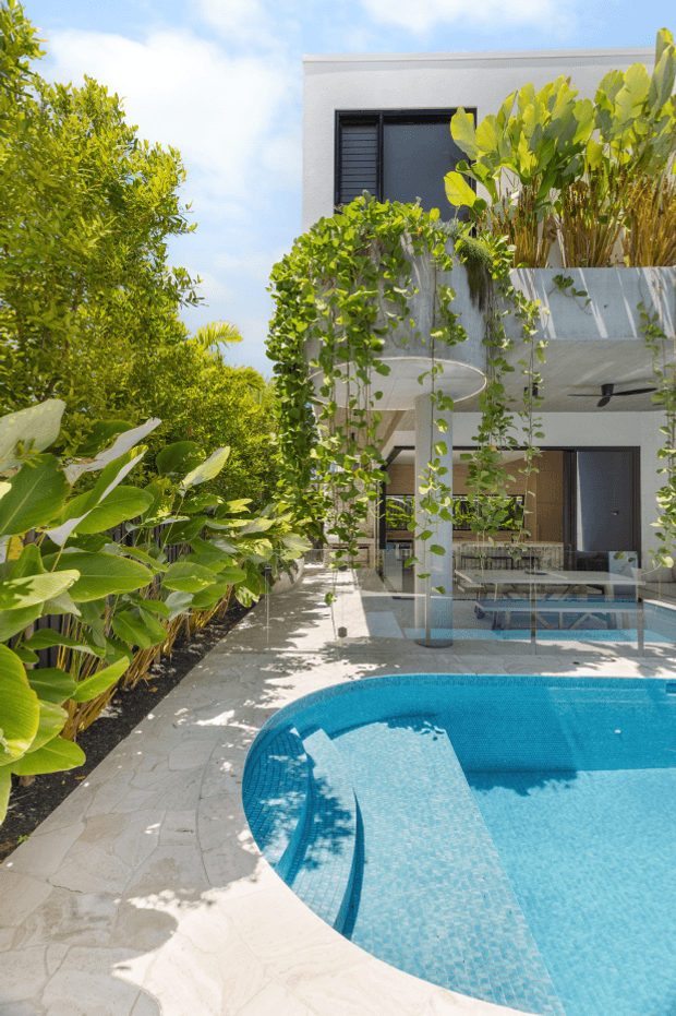 A bright tropical courtyard centered on a curved, turquoise-tiled swimming pool. Pale stone paving surrounds the pool, with dense lush planting, trailing vines, and tall broad-leaf plants framing a contemporary multi-storey white building. The building has dark-framed windows and doors, a projecting upper balcony or terrace with integrated planters, and a shaded ground-level outdoor area. The image has a sunny, resort-like atmosphere with strong greenery and water as visual features.