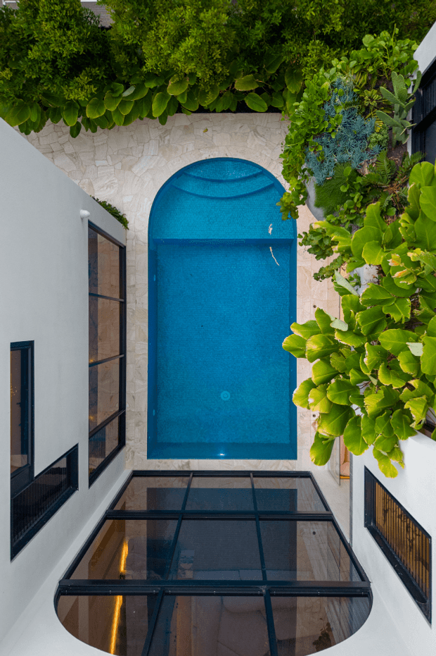 A top-down architectural view of a narrow courtyard with a vivid blue swimming pool, rounded pool end and steps, surrounded by pale stone paving, white rendered walls, black-framed glazing, and dense tropical planting. The enclosed courtyard composition emphasizes the contrast between the pool, modern building surfaces, and lush greenery.