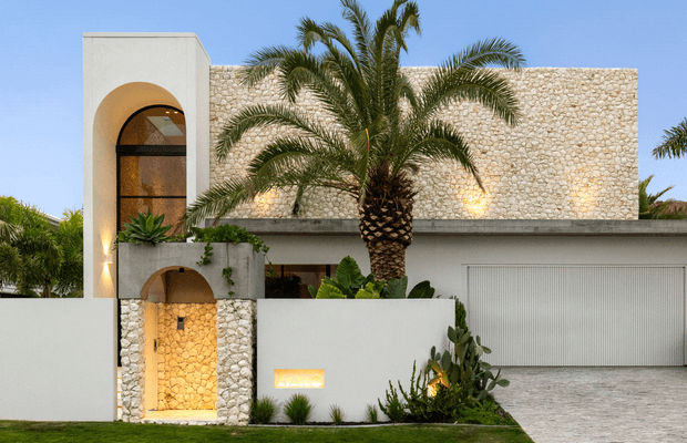 Front exterior view of a contemporary residence with a sculptural white stucco facade, arched entry and upper window, pebble-stone feature walls, a concealed garage door, and lush palm and tropical planting. Warm architectural lighting highlights the entry and textured masonry against a clear blue sky.