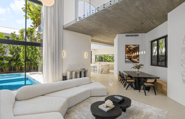 Bright, double-height contemporary living and dining area opening onto a courtyard swimming pool. The room features a large curved light fabric sectional, black round coffee tables, a black dining table with matching chairs, pale timber flooring, white walls, extensive glazing, and a dramatic exposed concrete ceiling with an upper-level balustrade. A kitchen is visible beyond the dining area.
