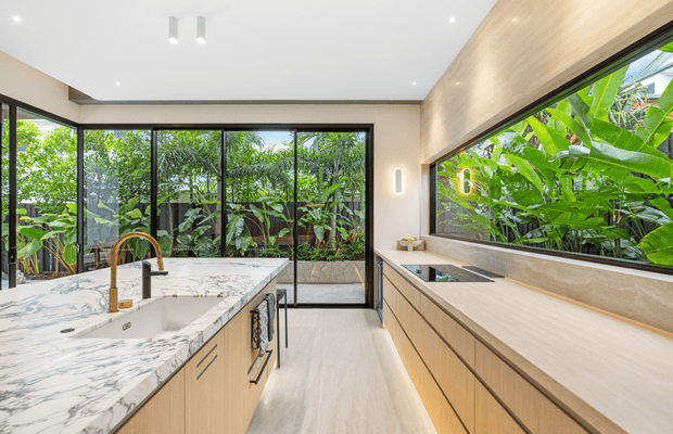 Contemporary kitchen with light timber cabinetry, a large white marble island, integrated appliances, and a long horizontal window overlooking lush tropical foliage. Full-height black-framed glazing brings abundant natural light into the space, complemented by discreet recessed and wall lighting.