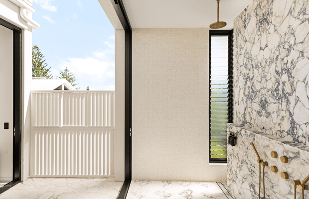 Bright contemporary bathroom with a walk-in shower and freestanding-style shower fixtures, featuring dramatic white-and-grey veined stone or marble-look wall cladding and matching floor tiles. A narrow louvred window provides daylight, while a white vertical-slat privacy screen is visible through the open glazing. Black-framed openings and brushed brass shower hardware add contrast.