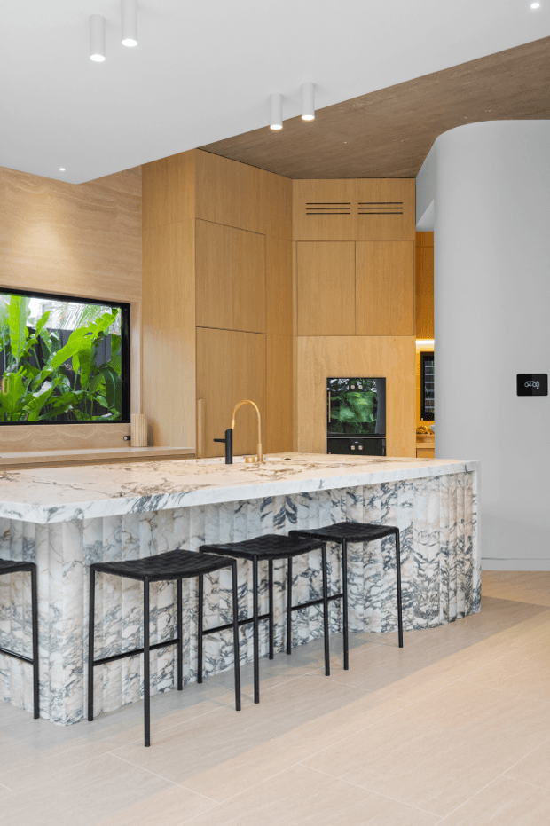 Bright contemporary kitchen with a large veined stone island and integrated seating, warm timber cabinetry and ceiling lining, a black-framed garden-facing window, built-in appliances, and minimalist white pendant and recessed lighting. Slim black bar stools line the island, while a curved white wall and pale large-format floor tiles complete the clean, refined interior.