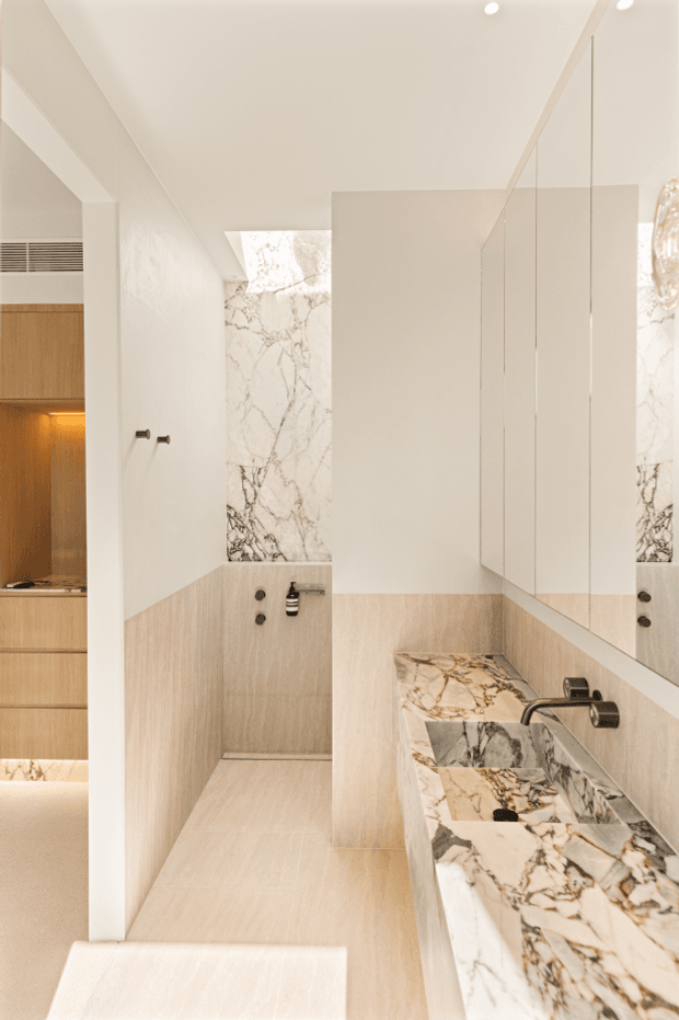 A refined contemporary bathroom with a walk-in shower, large mirrored wall, floating vanity, and sculptural marble-look stone basin and benchtop. The space uses warm pale timber-toned wall and floor finishes, white plaster, black tapware, and softly integrated lighting. A partial timber cabinetry zone is visible beyond the bathroom.