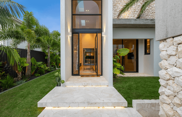 Contemporary residential entrance viewed from the landscaped front garden, featuring a tall glazed entry door with black metal framing, pale textured stucco walls, extensive irregular natural-stone cladding, and illuminated pale stone steps. Tropical palms and lush planting surround the entry, while warm interior lighting is visible through the glazing.
