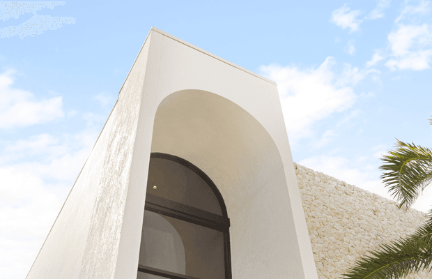 Low-angle exterior view of a sculptural white stucco facade featuring a tall recessed arched entry or window, with a contrasting pale rough-stone wall and palm fronds at the edge of the frame. The bright, geometric composition suggests a modern Mediterranean-inspired building exterior.