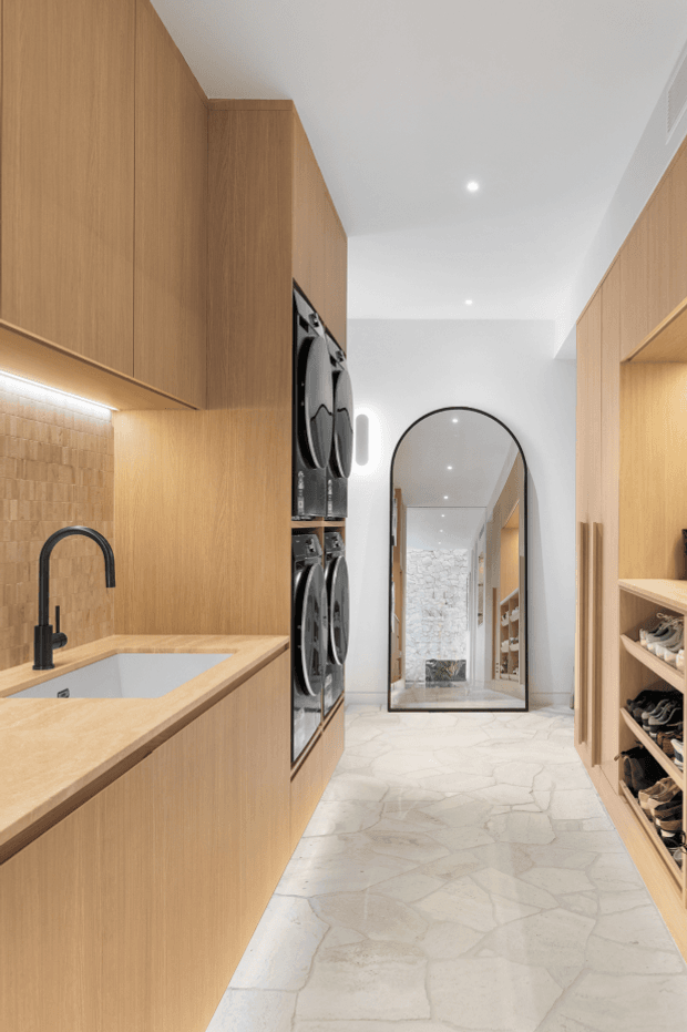 A narrow, contemporary utility and laundry corridor with warm light-wood cabinetry, integrated stacked washing machines, a timber-toned benchtop with inset sink and matte-black gooseneck tap, open shoe storage, and pale irregular stone-look flooring. A tall arched mirror at the end reflects the hallway and a textured stone feature wall.