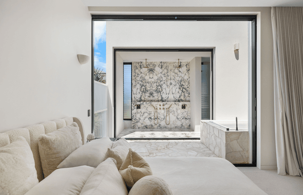 Bright, luxurious bedroom opening directly onto a marble-clad ensuite bathroom. The foreground features a large cream upholstered bed with plush pillows, while black-framed glazing and curtains surround the room. The ensuite includes a freestanding bathtub, double vanity, and extensive veined marble wall and floor surfaces.