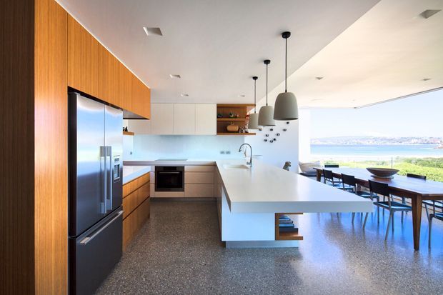 Bright open-plan kitchen and dining area with a large white island, integrated stainless-steel appliances, warm timber cabinetry, terrazzo flooring, pendant lights, and expansive glazing overlooking a coastal landscape.