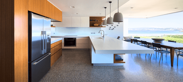 A bright contemporary open-plan kitchen and dining area with extensive warm timber cabinetry, a large white island bench, dark pendant lights, integrated stainless-steel appliances, and a polished terrazzo-like floor. Full-height glazing opens the room to a waterfront or coastal view, with a dining table positioned beside the windows.