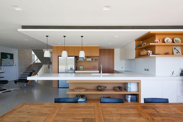 Open-plan contemporary kitchen and dining area with a large white island, integrated stainless-steel appliances, warm timber cabinetry and open shelving, pendant lights, and a substantial rustic timber dining table. A stairway and living area are visible in the background.