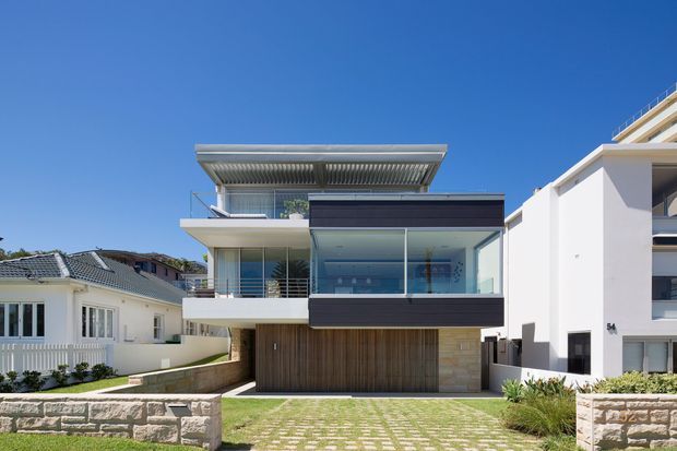 Exterior street-facing view of a contemporary multi-level residence with a flat roof, cantilevered upper terrace, extensive glazing, a projecting dark-framed glass volume, vertical timber cladding, pale masonry walls, and a landscaped permeable-paver driveway. Adjacent homes are visible on either side under a clear blue sky.
