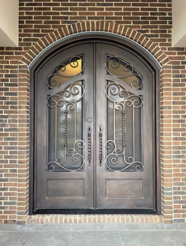 Close-up exterior view of an ornate arched double entry door set within a dark red brick facade. The dark metal doors feature decorative scrollwork, inset glass panels, elongated metal handles, and a brick voussoir arch above, with stone paving at the threshold.