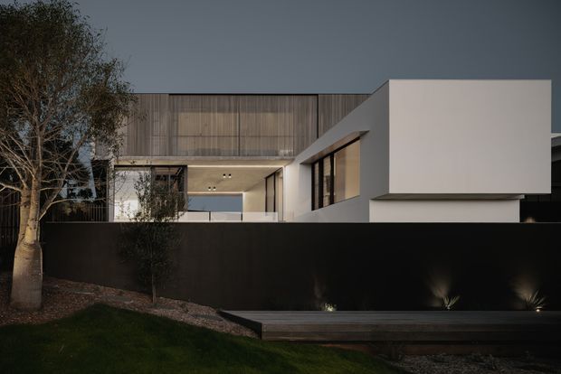 A contemporary multi-level residence viewed from the street at dusk. The home features crisp white rendered volumes, extensive dark-framed glazing, warm vertical timber screening, and a flat roofline. A tall matte-black boundary wall with subtle landscape lighting encloses a timber-edged front terrace, with mature trees and planting in the foreground.