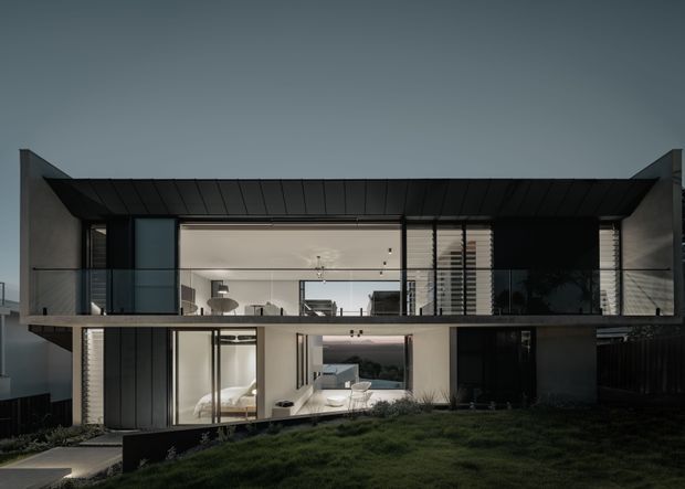A contemporary two-storey residential home photographed externally at dusk. The dark, geometric facade features a broad projecting upper level, extensive floor-to-ceiling glazing, slim black window frames, glass balustrades, and a shallow pitched or folded metal roof form. Warmly lit living spaces are visible through the glazing, with landscaped lawn and low garden planting in the foreground.
