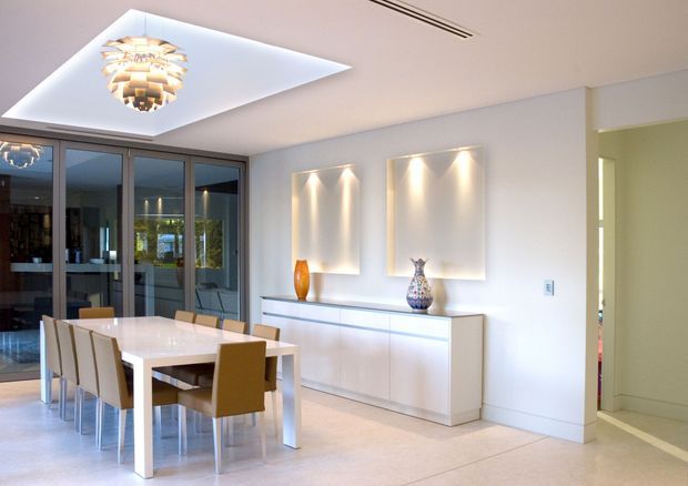 Bright contemporary dining room with a long white dining table and upholstered chairs, a streamlined white sideboard, illuminated abstract wall panels, and a sculptural pendant light set into a recessed ceiling feature. Full-height dark-framed glazing opens toward an adjoining outdoor or living area, while a hallway is visible at right.