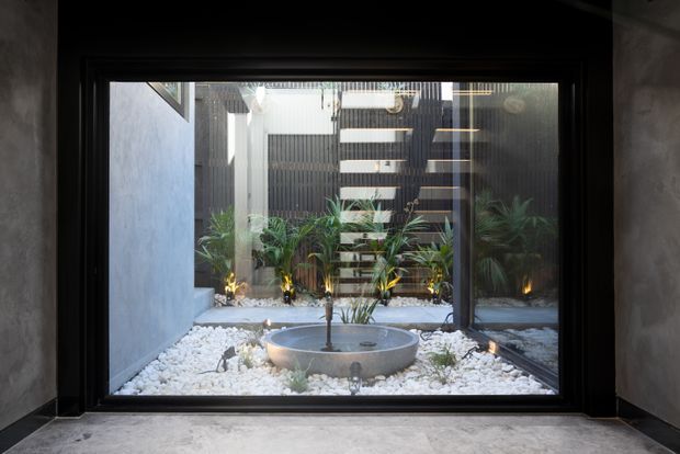 A dark, refined interior viewpoint looks through a large black-framed glazed opening onto a compact landscaped courtyard. White pebbles surround a shallow circular stone water feature, with tropical planting, architectural lighting, and a dark vertical screen beyond. The interior has smooth grey plaster walls and a pale concrete-like floor, creating a calm contemporary transition between indoors and outdoors.