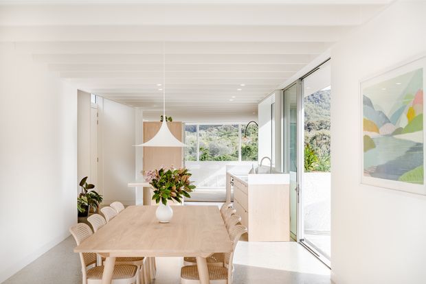 Bright, open-plan dining area with a long light timber dining table and woven dining chairs, connected to a compact kitchen through full-height glazed doors. White walls and exposed white ceiling beams create an airy interior, with a sculptural pendant light, pale cabinetry, indoor planting, and artwork. Greenery and a hillside are visible beyond the glazing.