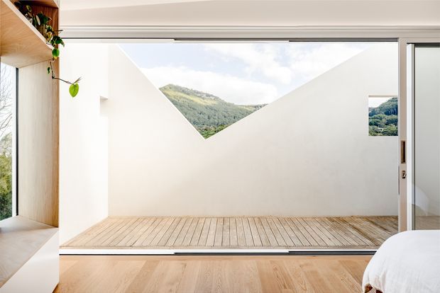 Bright minimalist bedroom opening onto a timber-decked courtyard or terrace. Full-width sliding glazing frames a sculptural white wall with a triangular mountain view, while pale timber flooring and built-in timber shelving create a warm, calm interior.
