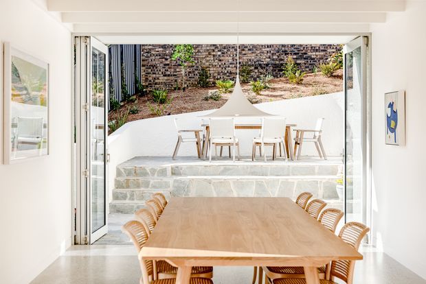 Bright contemporary dining room opening through fully glazed folding doors onto a raised outdoor dining terrace and landscaped courtyard. The room features a large light timber dining table with woven timber chairs, white walls, pale stone steps and retaining walls, and strong indoor-outdoor visual connection.