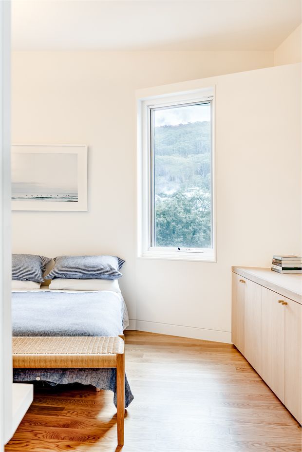 Bright, compact bedroom with a neatly made bed, striped blue-grey bedding, pale walls, timber flooring, a light timber built-in cabinet, and a tall white-framed window overlooking greenery. A simple coastal artwork and woven bench add understated natural texture.