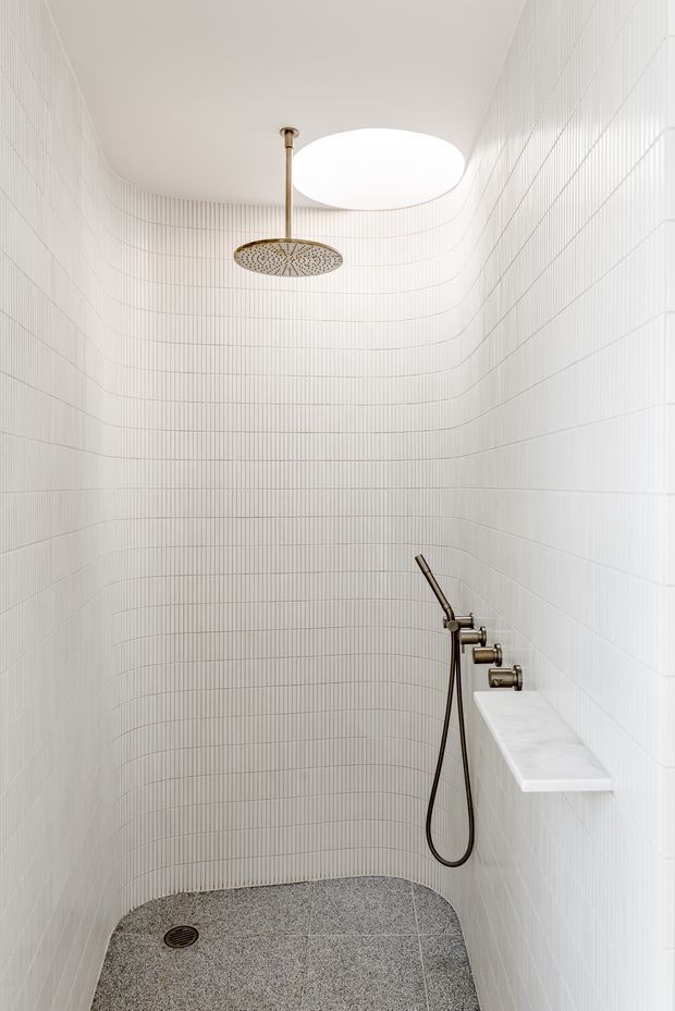 A compact contemporary walk-in shower with curved white vertically ribbed wall tiles, a white ceiling with a large circular skylight or recessed light, and brushed brass shower fittings. The floor is finished in grey speckled stone or terrazzo tile, with a built-in white ledge and handheld shower.
