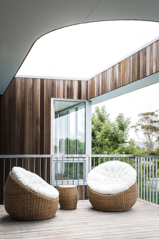 A covered outdoor terrace or balcony with a timber deck, two large rounded woven lounge chairs with white cushions, and a small woven side table. The space is enclosed by silver metal balustrades and framed by dark vertical timber wall cladding, with a large open skylight-like roof opening and expansive glazing/door. Trees and a distant urban landscape are visible beyond the railing.