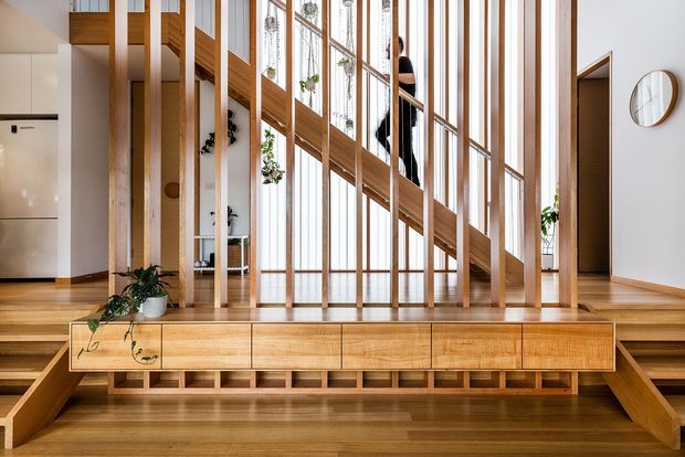 A bright, open-plan residential interior centred on a sculptural timber staircase and mezzanine. Vertical timber posts and large glazed panels create a light-filled stair hall, with built-in timber cabinetry and open shelving beneath the stairs. Warm timber flooring, white walls, indoor plants, and minimal furnishings create a refined contemporary atmosphere. A kitchen area is visible at left and a hallway or doorway at right.