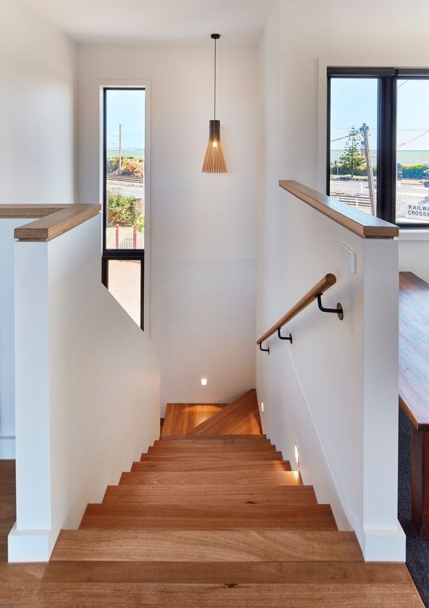 Bright interior photograph looking down a timber staircase. The stair treads, upper landing handrails, and balustrade caps are warm natural timber, contrasted with crisp white walls and stair sides. Black-framed windows and a narrow glazed door bring in abundant daylight and show a coastal suburban street beyond. A simple timber pendant light hangs over the stairwell, while a timber handrail with dark metal brackets runs along the wall.