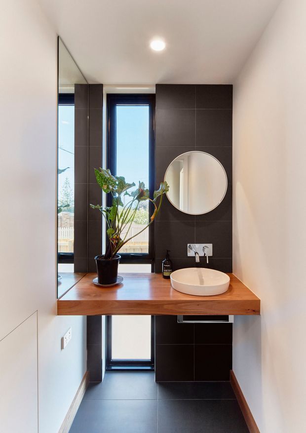 Compact contemporary bathroom vanity area with a wall-mounted timber countertop, white vessel basin, round mirror, dark tiled feature wall, black-framed windows, and integrated dark cabinetry. A large mirror reflects the bright narrow room, while a potted plant adds natural contrast.