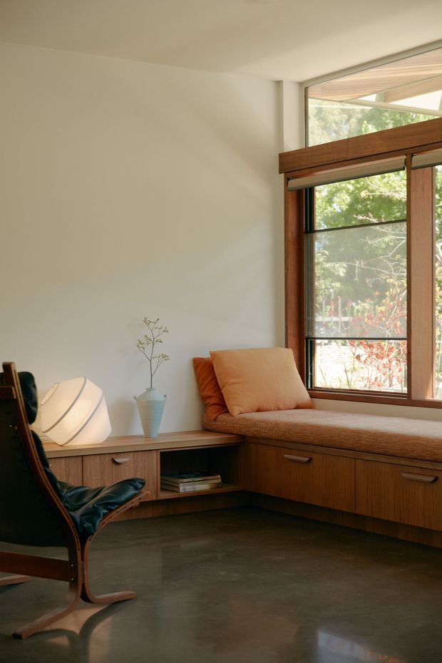 A warm, mid-century-inspired reading nook with a built-in timber window seat and low storage cabinetry beneath a large timber-framed window. The space includes a dark leather lounge chair, a softly glowing sculptural lamp, a blue ceramic vase, and a neutral upholstered cushion. Smooth polished concrete flooring and off-white walls create a calm, understated interior, with greenery visible outside the glazing.