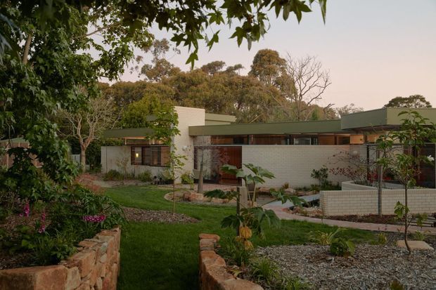 Single-storey low-profile residence set within a landscaped garden, viewed from the front. The home has long horizontal proportions, pale masonry walls, dark green flat roof planes, timber-framed glazing, and sheltered outdoor circulation. Natural stone retaining walls, gravel landscaping, lawn, and mature trees surround the building, giving the scene an integrated garden setting.