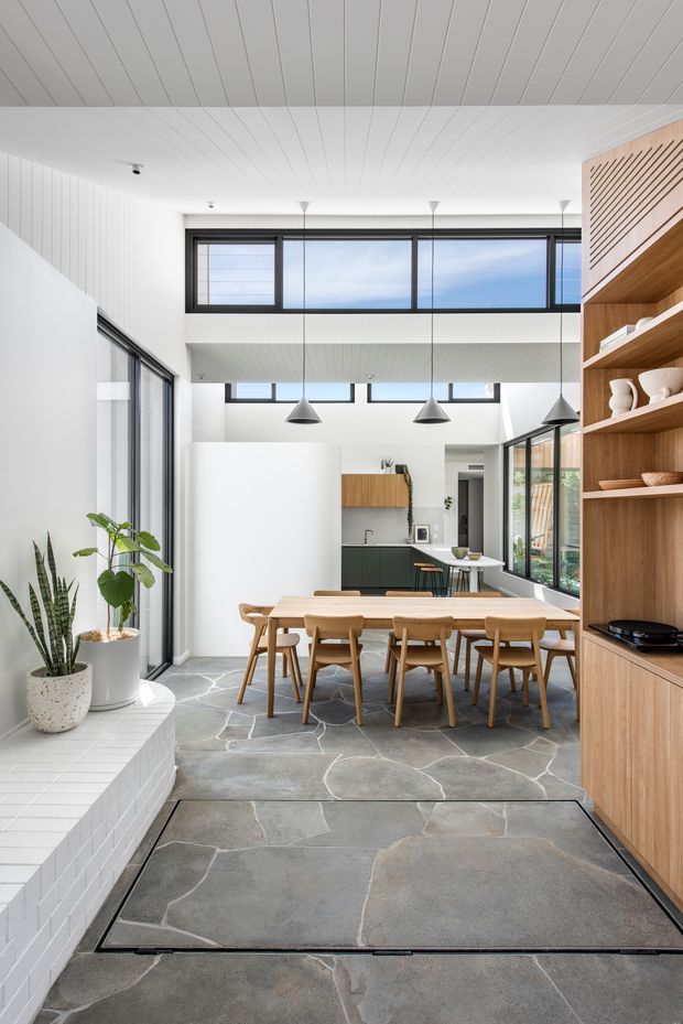 Bright open-plan dining and kitchen interior with a double-height ceiling and clerestory glazing. A large timber dining table and chairs occupy the foreground, with built-in timber shelving and cabinetry on the right, a compact kitchen at the rear, pendant lights, white-painted surfaces, dark-framed windows, indoor plants, and irregular grey stone-look flooring. The space feels airy, contemporary, and connected to the outdoors.